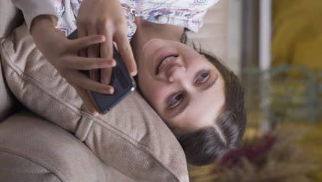 Vertical-video-of-Young-woman-lying-on-sofa-is-using-phone.-Pleasant-and-comfortable-posture.